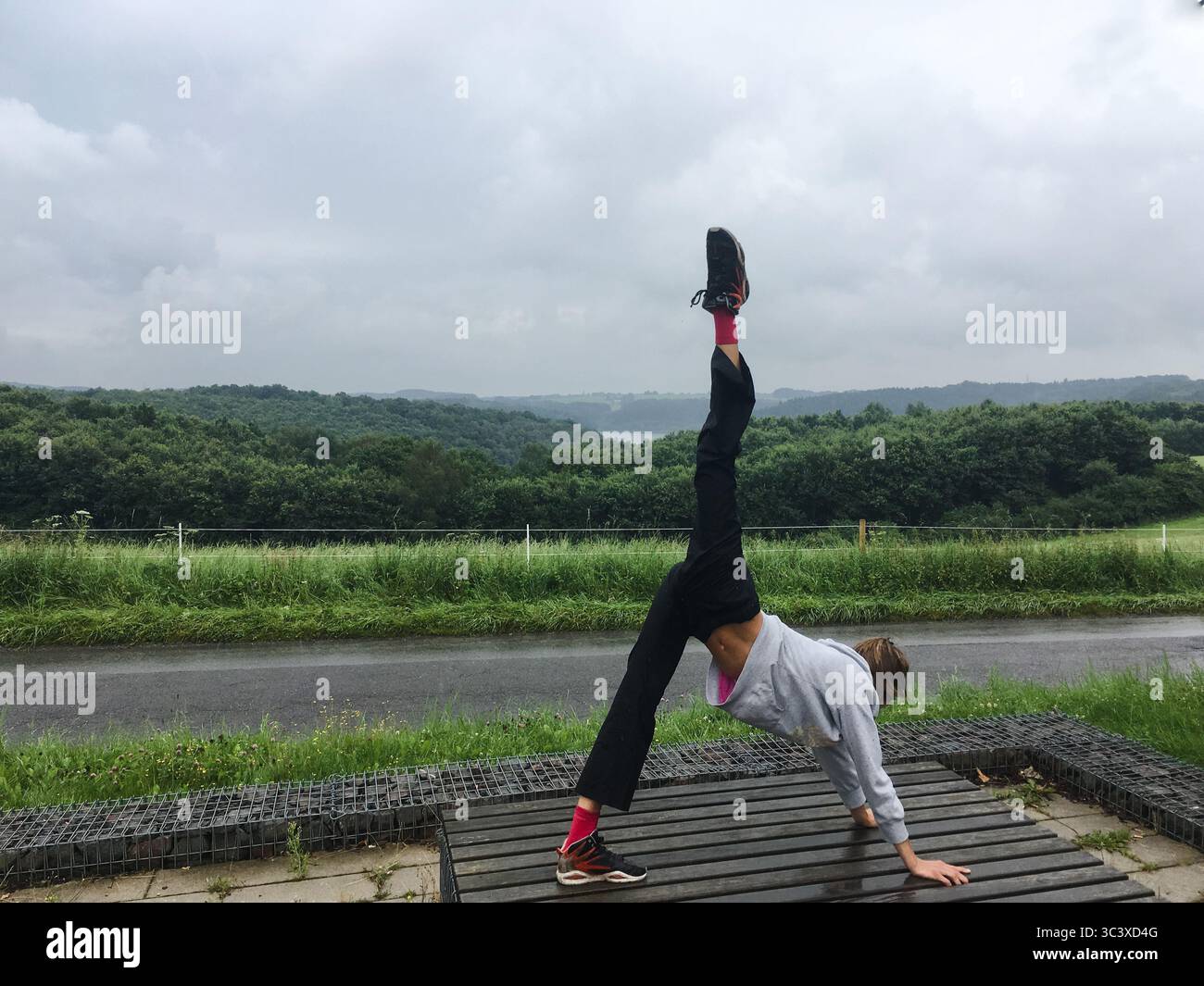 Una giovane donna irriconoscibile che fa yoga con cane a tre zampe si posa quasi come una spaccata di fronte all'ambiente verde della natura. Stile di vita attivo, yoga. Foto Stock