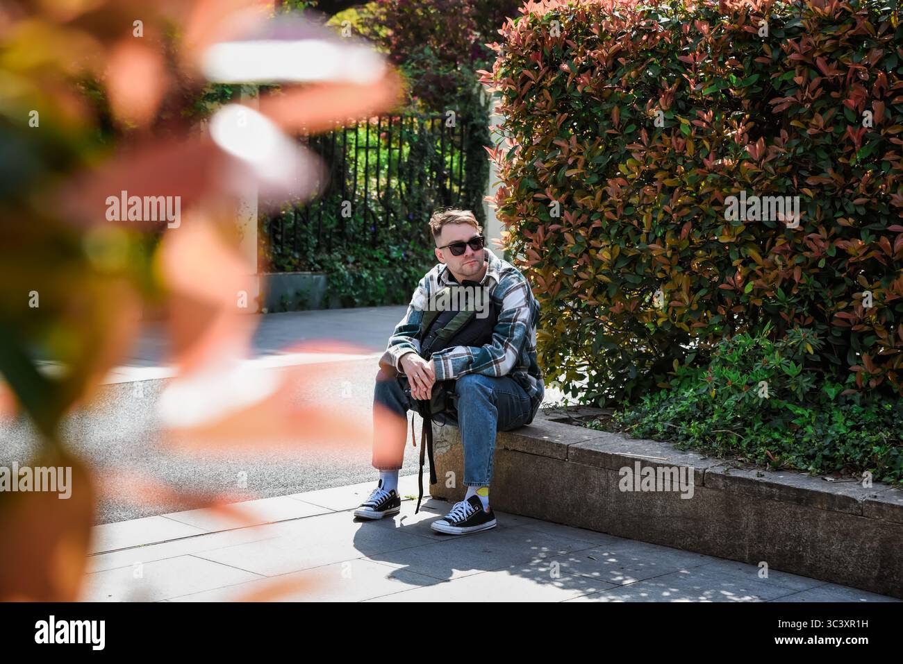 Un turista russo maschio che riposa su una panchina di pietra vicino agli arbusti autunnali in un parco cittadino, godendo di una giornata di sole durante i suoi viaggi. Foto Stock