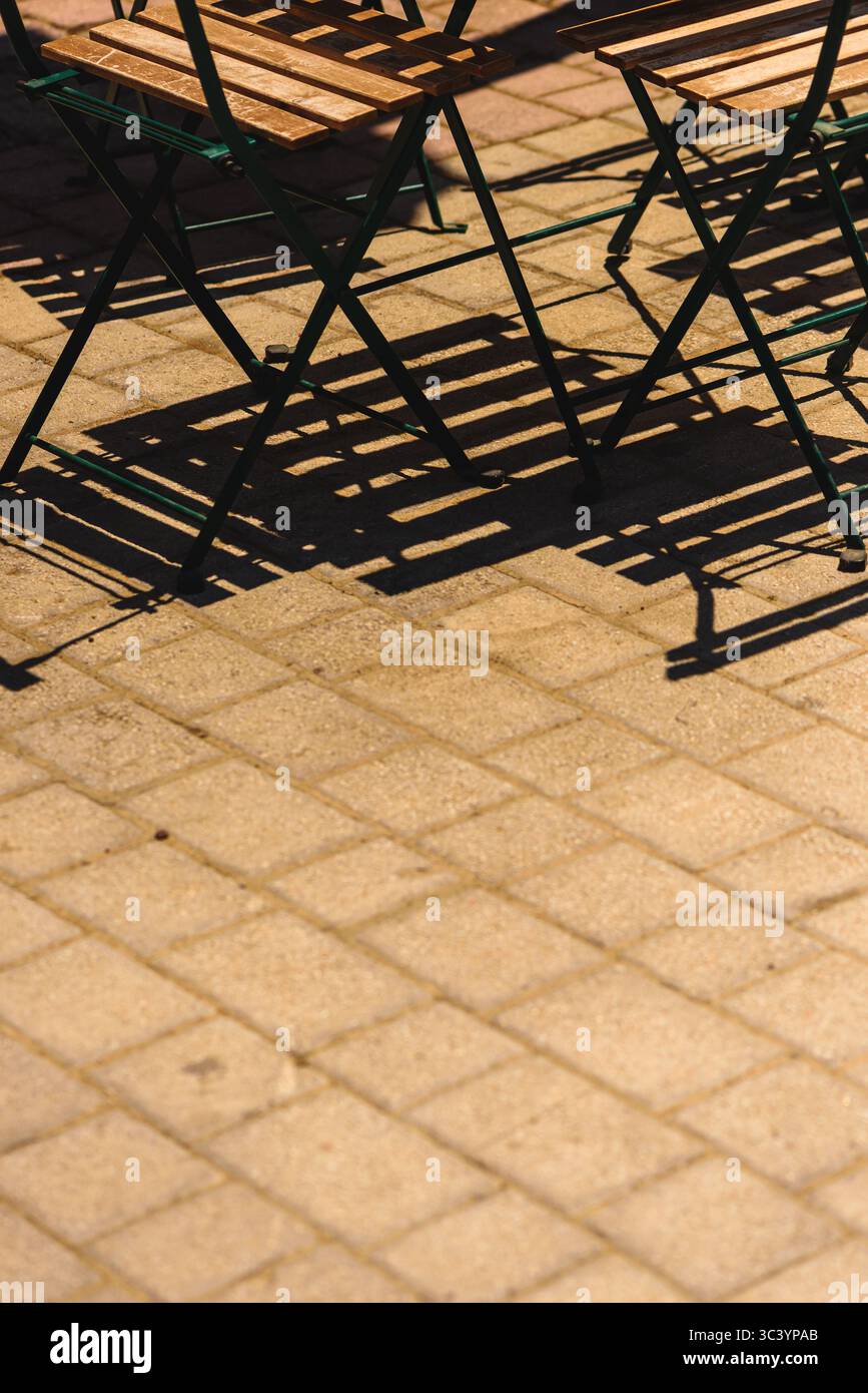 Ombre di sedie vuote in metallo e legno gettate su una pavimentazione piastrellata illuminata dal sole in un ambiente caffetteria all'aperto. Messa a fuoco selettiva. Foto Stock