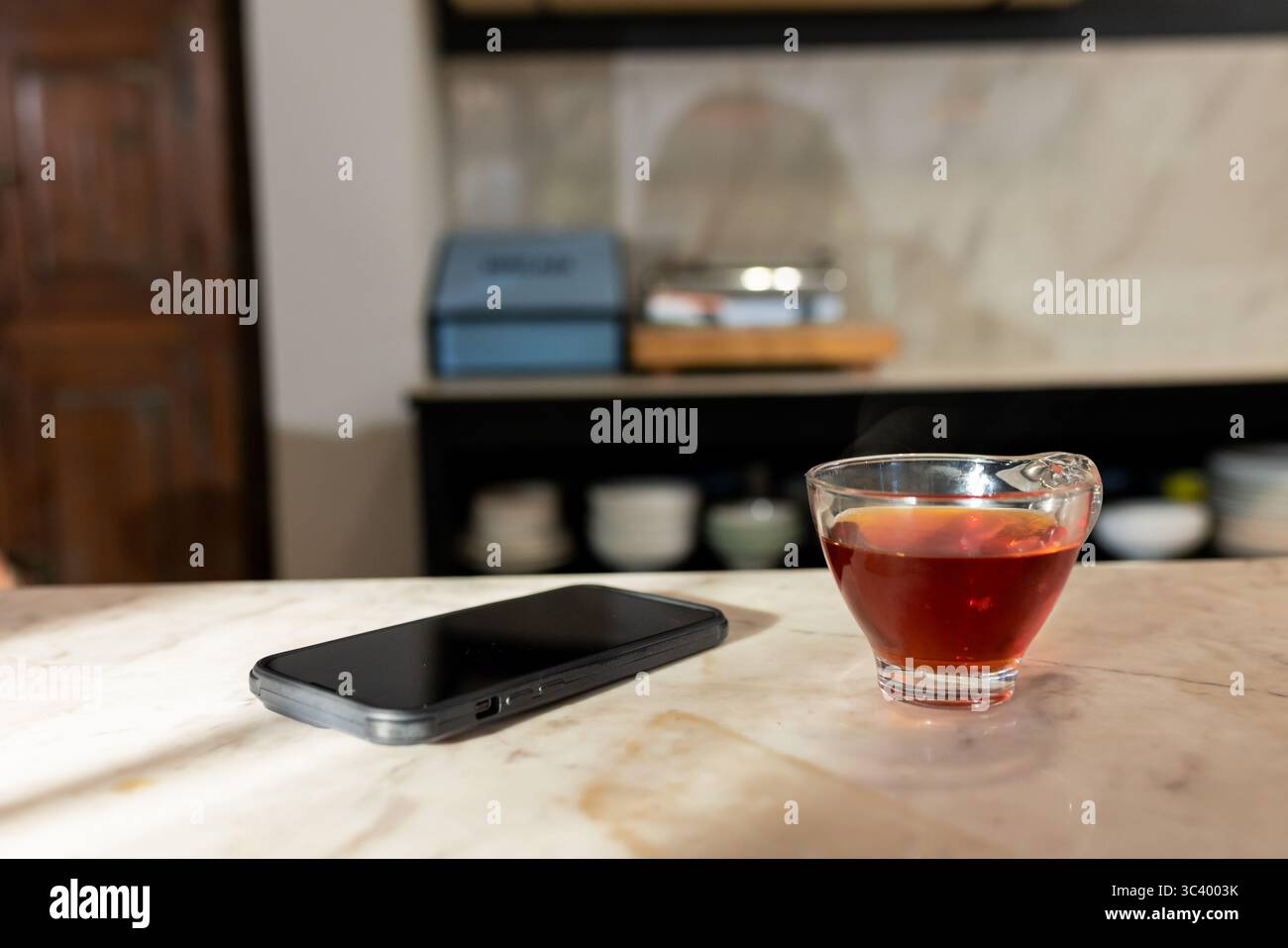 Smartphone adagiato accanto a una tazza di tè scuro su un piano di appoggio in marmo, con ciotole e scatole sfocate dietro Foto Stock