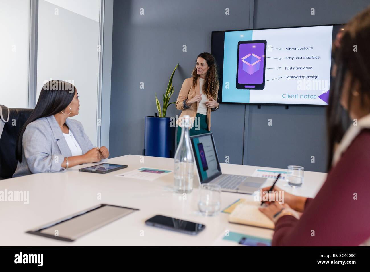 Donne d'affari diverse che presentano diapositive con interfaccia mobile al tavolo da conferenza con notebook e tablet Foto Stock