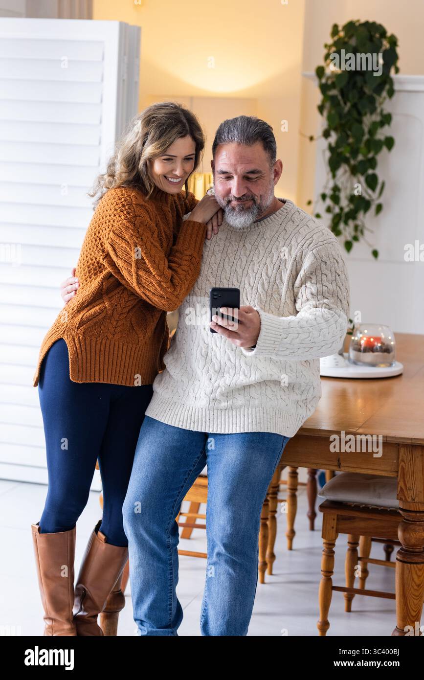 Coppia che indossa maglioni in maglia a cavo appoggiati al tavolo da pranzo guardando lo smartphone vicino alla pianta in vaso Foto Stock