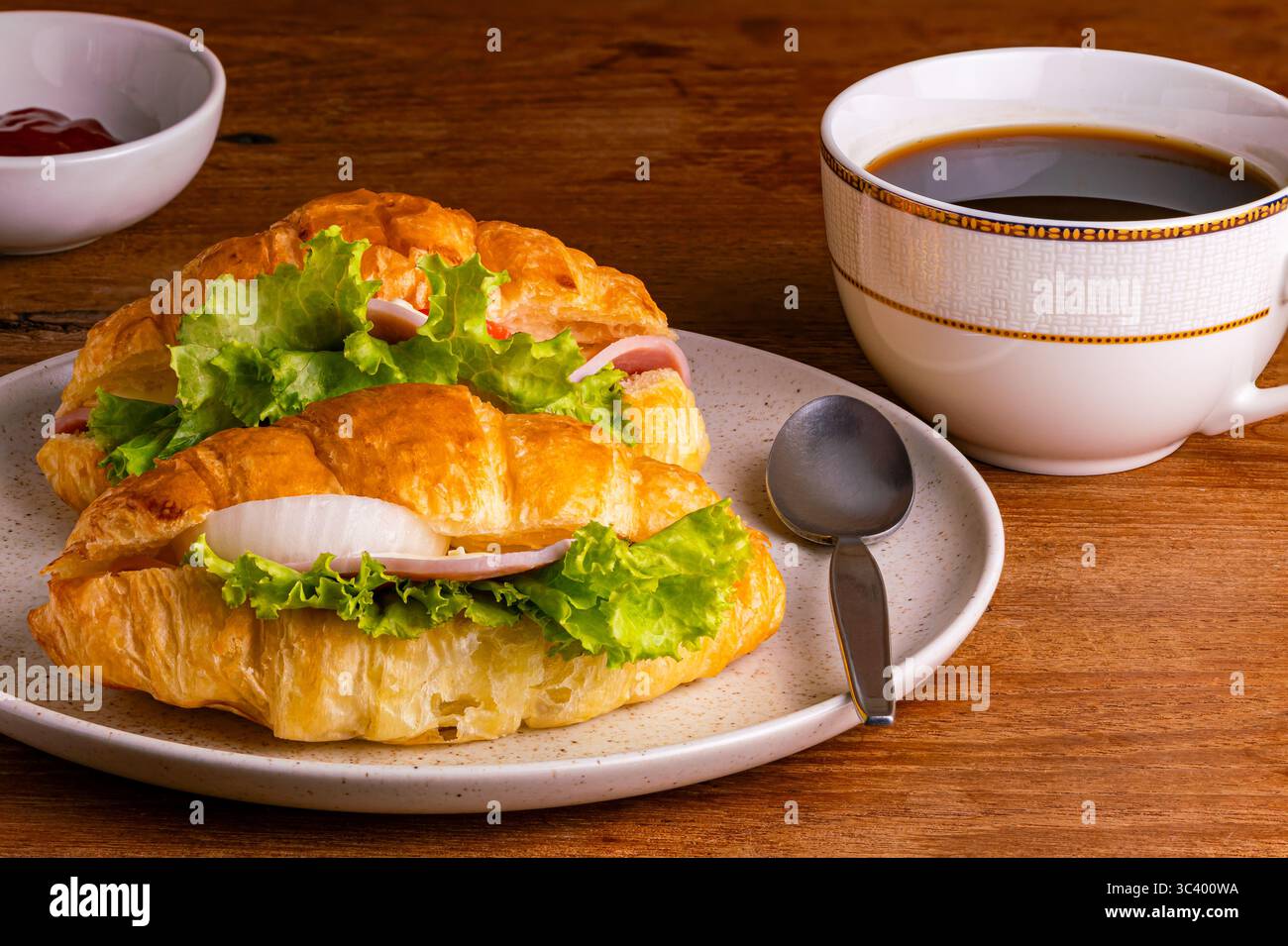 Croissant prosciutto con verdure verdi e cipolla in un piatto di ceramica macchiata marrone con una tazza di caffè nero e ketchub in un fiocco di ceramica bianca Foto Stock