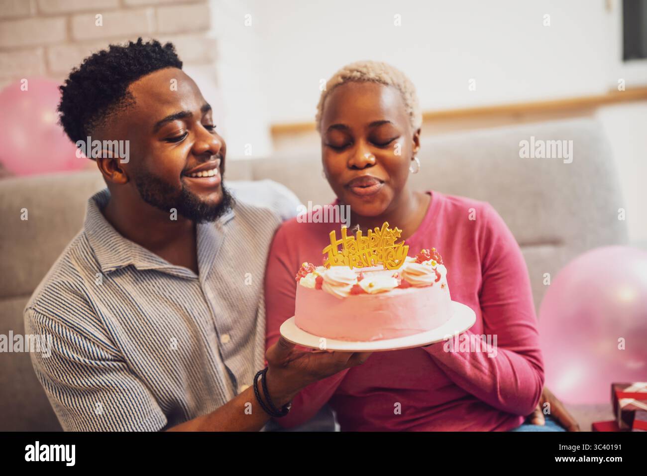 Felice giovane coppia che festeggia il compleanno della donna a casa. Stanno tenendo la torta e stanno soffiando la candela di compleanno. Foto Stock
