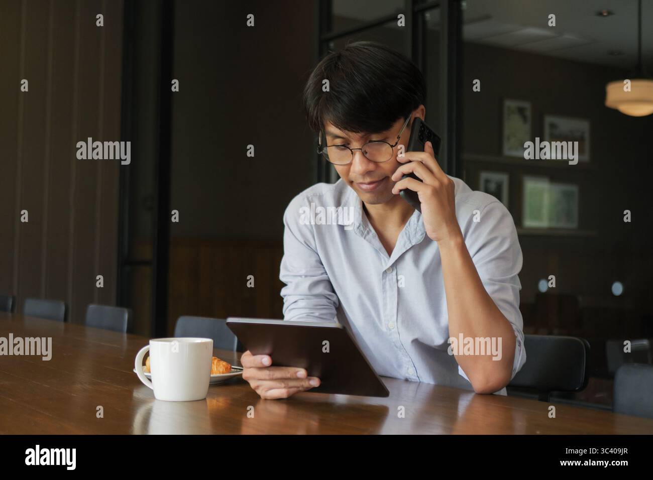 Uomo asiatico che indossa occhiali che parla al telefono, guarda le informazioni sui dati sul tablet, lavora al mattino sul tavolo da pranzo in legno con caffè caldo Foto Stock