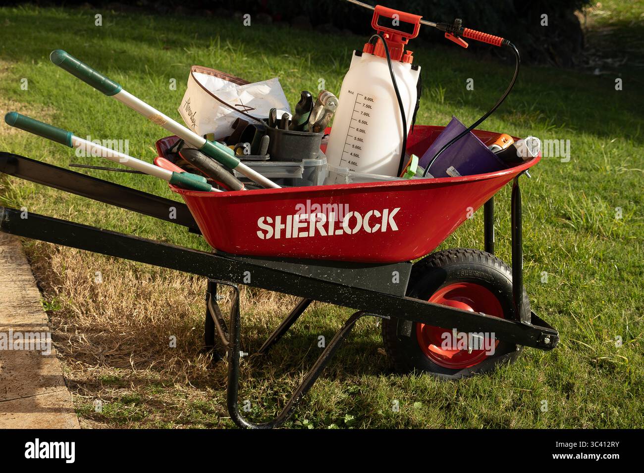 Carriola Sherlock in acciaio rosso brillante piena di attrezzi e spray seduti sul prato, pronti per essere utilizzati in un giardino nel Queensland, Australia. Foto Stock