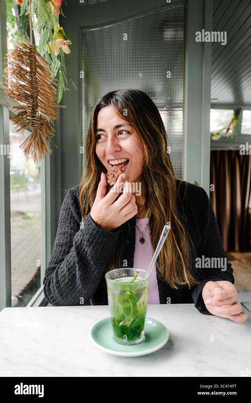 Donna sorridente che mangia un biscotto a un tavolo vicino alla finestra nel caffè Alkmaar Foto Stock