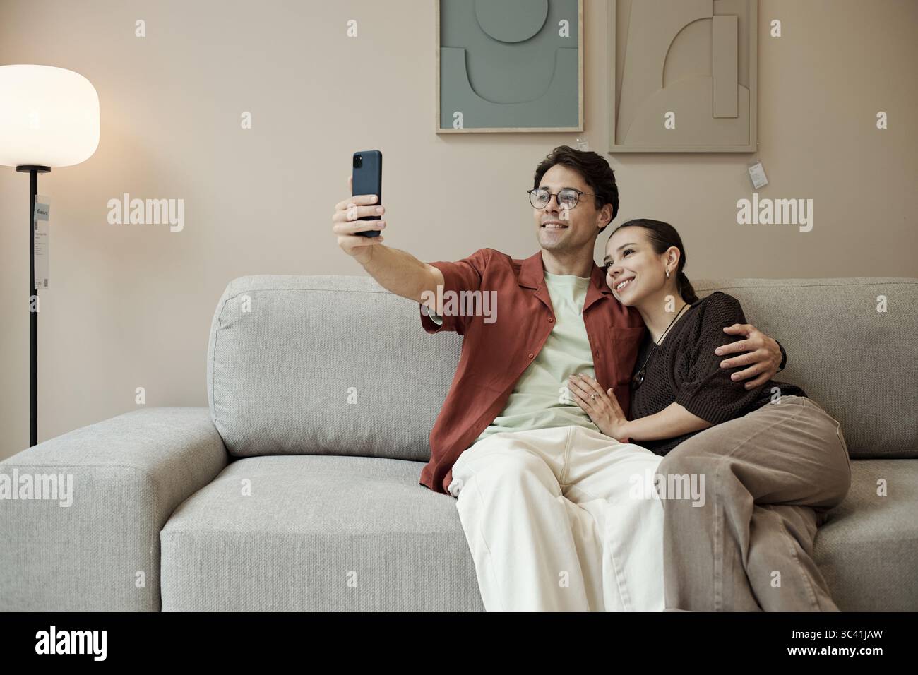 Giovane uomo caucasico adulto seduto sul divano a scattare selfie con lo smartphone, mentre la giovane donna caucasica adulta seduta accanto a lui abbraccia e sorride nello showroom del negozio di mobili Foto Stock