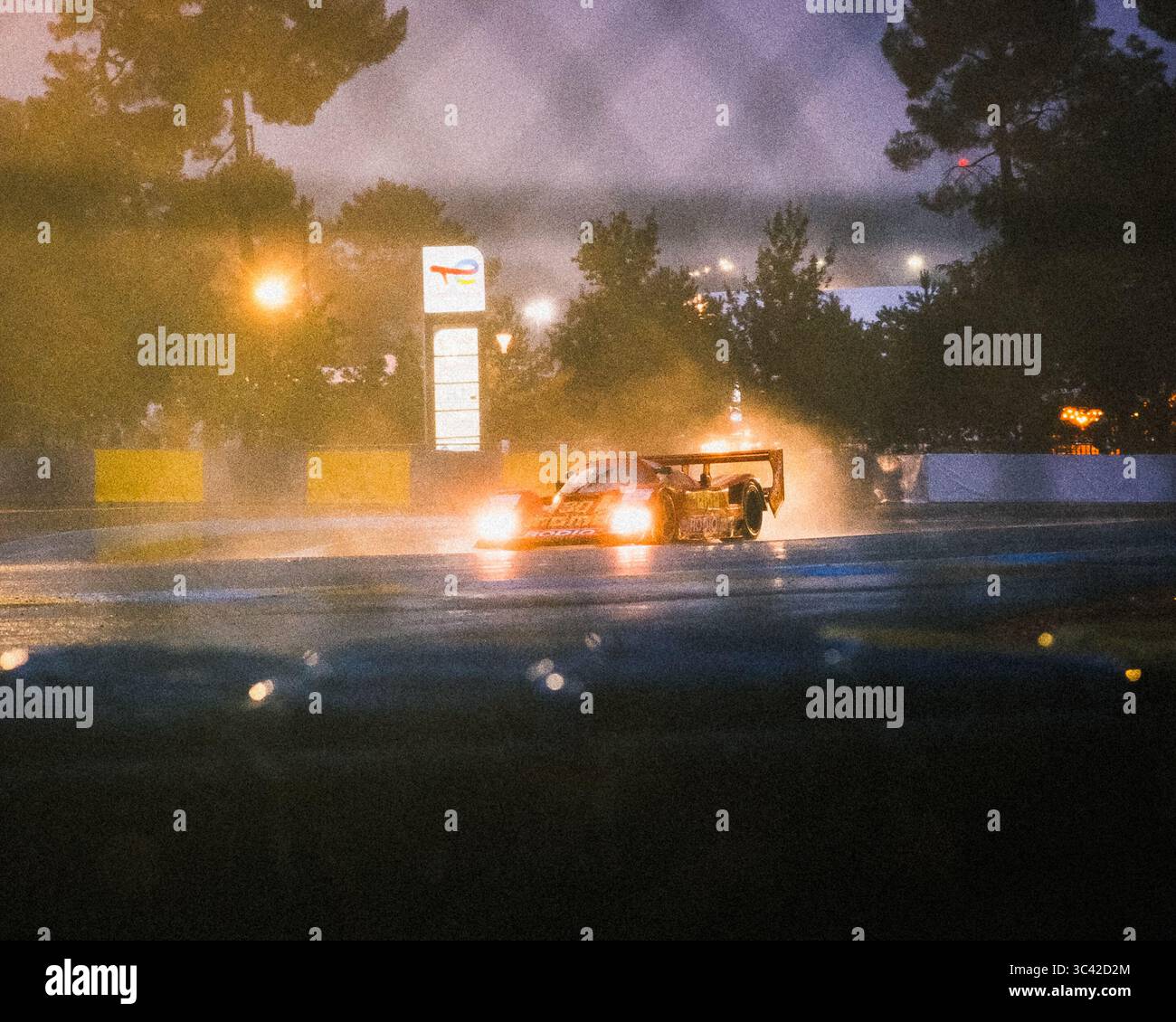Le Mans, Francia - 07 giugno 2025: Veduta di una vettura da corsa che accelera di notte attraverso Place Luigi Chinetti, illuminata da luci accese, con pioggia che si riflette sulla pista. Foto Stock