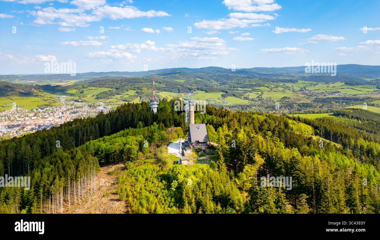 Una vista aerea cattura la torre di osservazione di Svatobor circondata da foreste lussureggianti e colline ondulate in Cechia. Il paesaggio sfoggia un mix di verde e montagne lontane sotto un cielo azzurro. Foto Stock