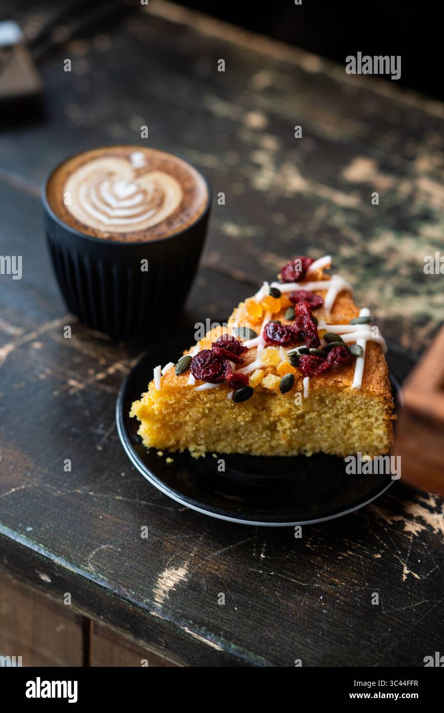 Una fetta di torta servita con una tazza di cappuccino con latte art, posta su un rustico tavolo di legno. Ambiente accogliente e invitante, perfetto per un ristorante Foto Stock