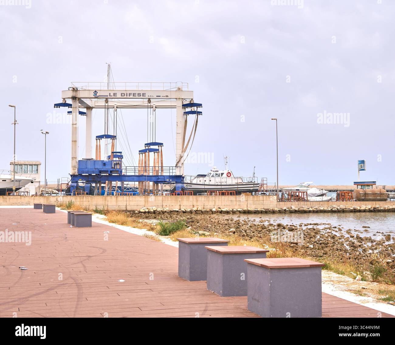 Cantiere marino con un grande ascensore, barche attraccate e un passaggio pedonale sul lungomare sotto un cielo nuvoloso Foto Stock