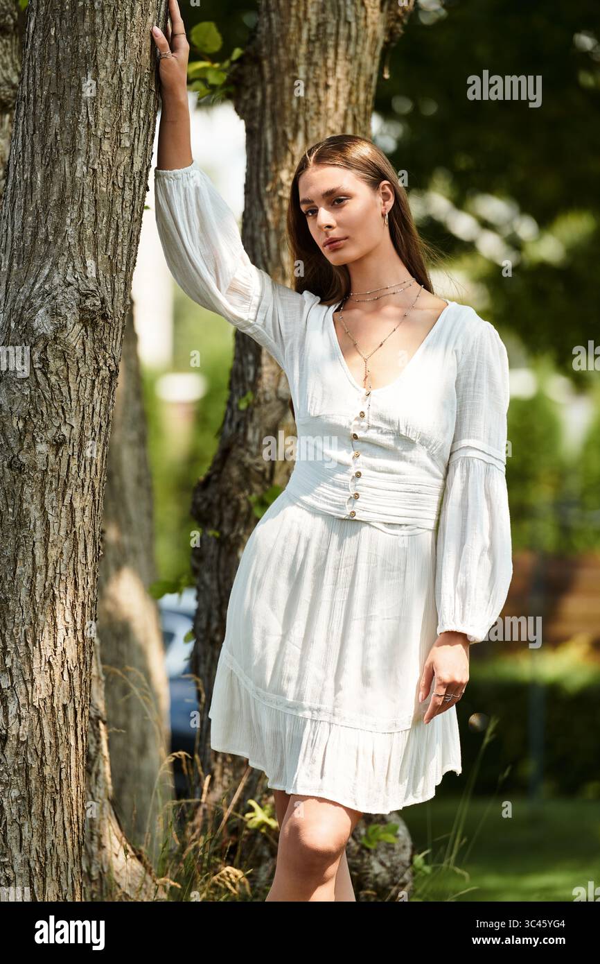 Una giovane donna posa elegantemente in un affascinante vestito bianco vicino a un albero, crogiolandosi alla luce calda del sole. Foto Stock