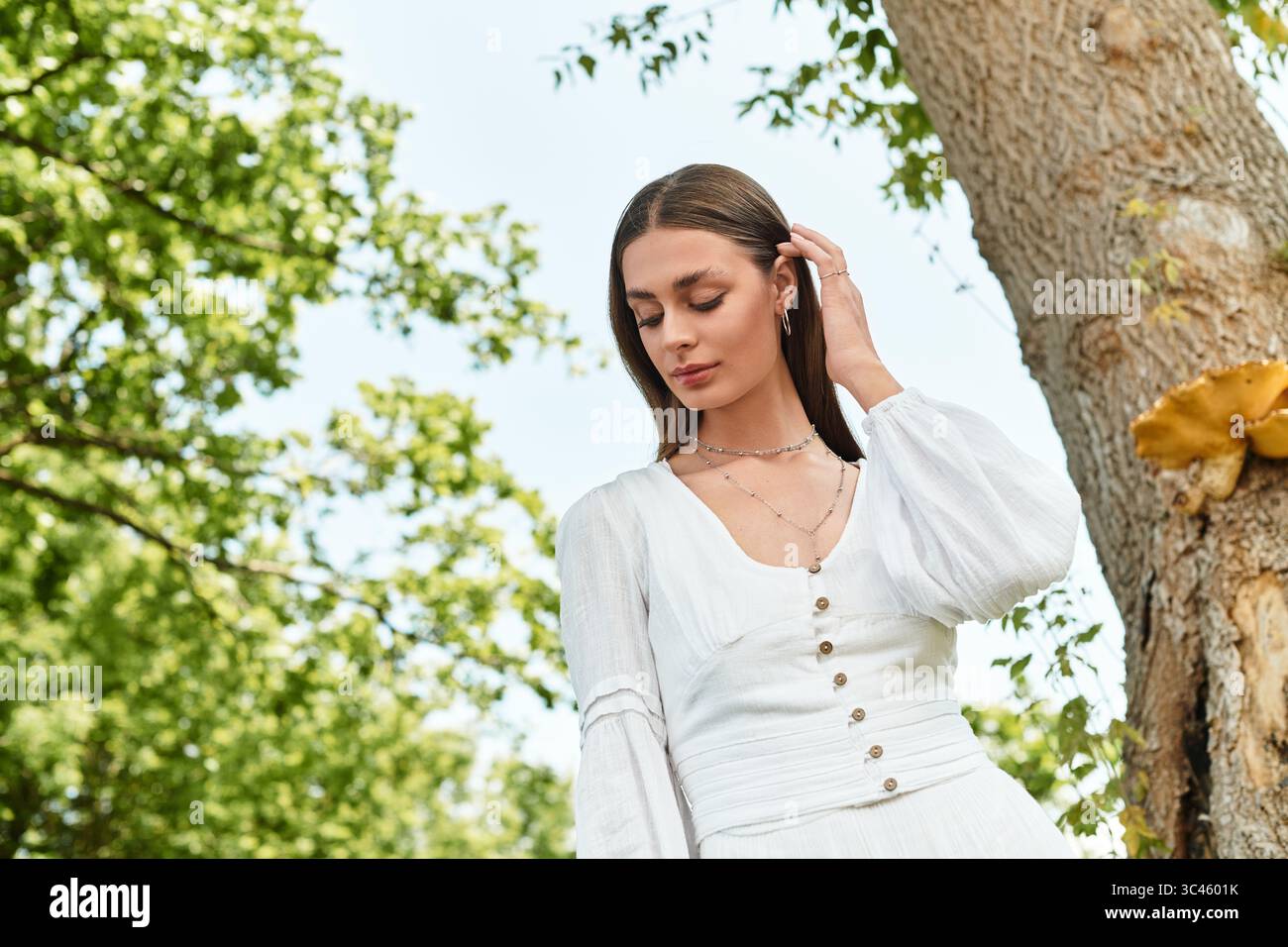Giovane donna che tocca delicatamente i capelli, perduta nel pensiero tra alberi vibranti in una giornata di sole Foto Stock