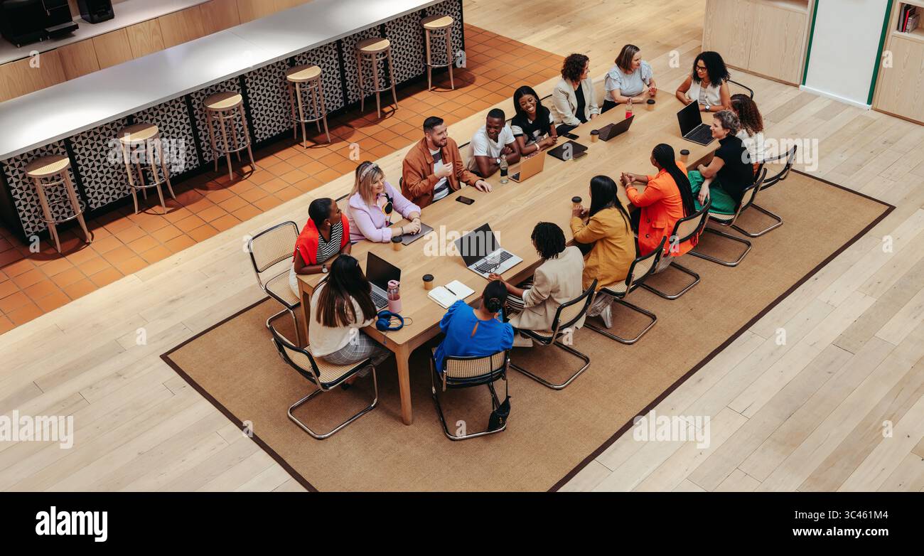 Una visione ad alto angolo di un gruppo eterogeneo di professionisti impegnati in una riunione di lavoro. L'ambiente è un ufficio moderno con un grande tavolo e computer portatili Foto Stock