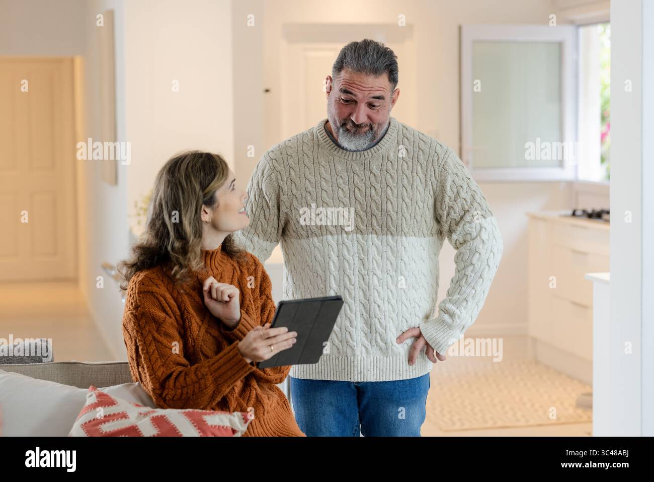 Abbina maglioni in maglia via cavo a discutere di tablet mentre sei seduto sul divano con un cuscino vicino alla cucina Foto Stock