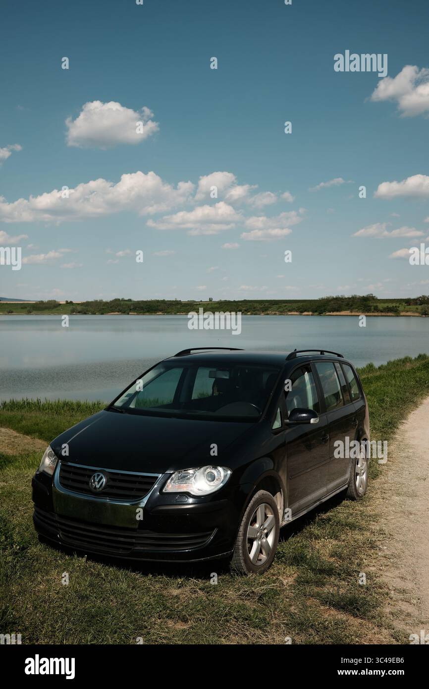 2 maggio 2025 - Lago Pavlovac, Serbia. Volkswagen Touran nero parcheggiato sotto un cielo blu brillante con nuvole soffici. Tranquillo paesaggio rurale. Viaggi, natura Foto Stock