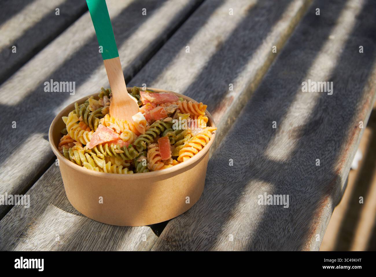 un'insalata di pasta colorata viene servita in una ciotola di carta marrone da asporto con un cucchiaio di legno, appoggiata su un tavolo da picnic intempestivo all'ombra con ombre di un Foto Stock