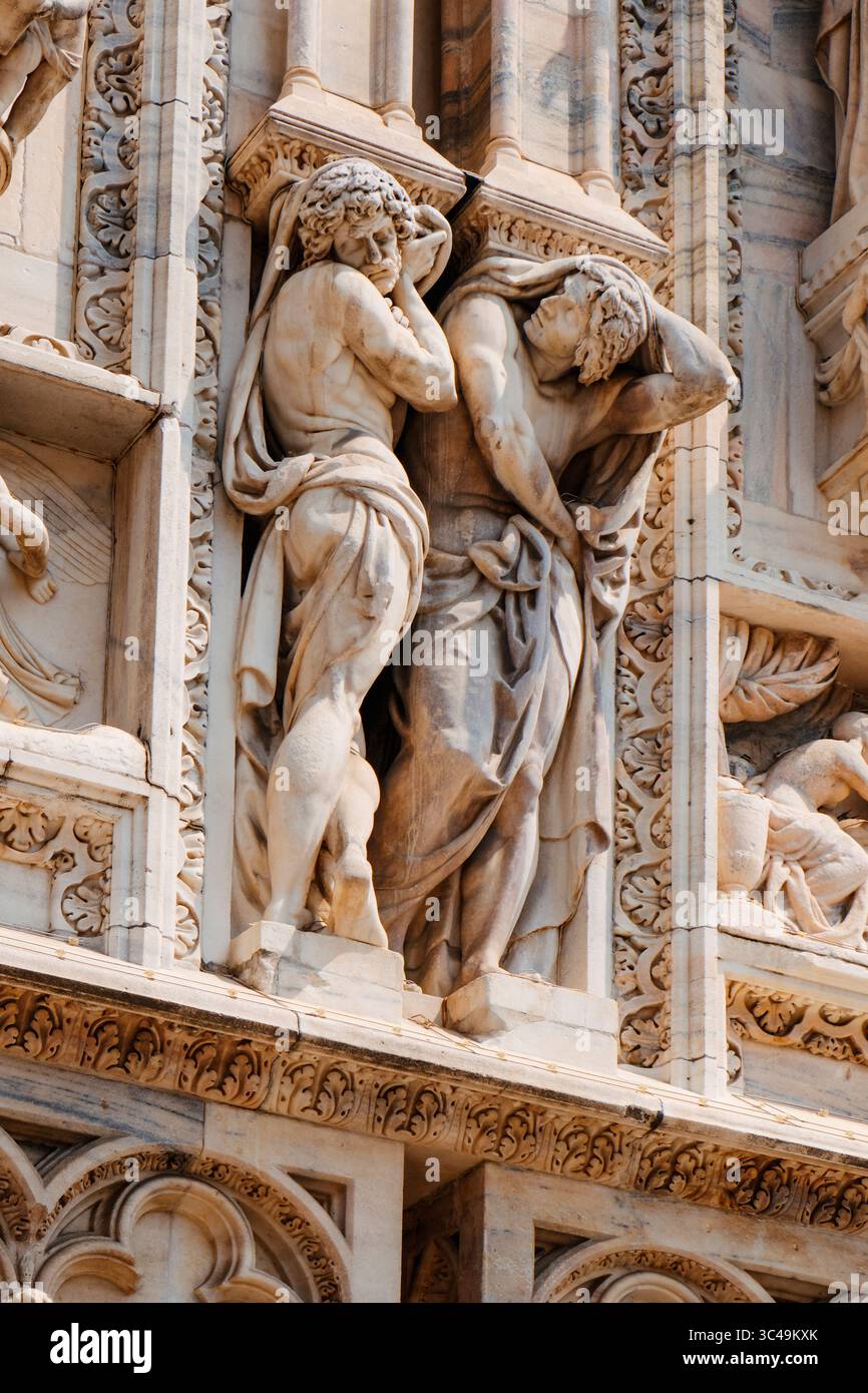 Due figure maschili scolpite sembrano sollecitare sotto il peso architettonico di un rilievo nella parte inferiore destra della facciata del Duomo di Milano Foto Stock
