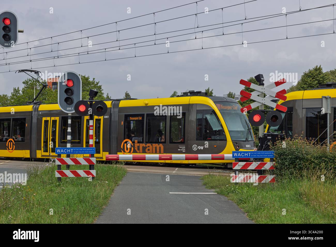 Due moderni tram gialli e grigi passano attraverso un passaggio ferroviario con barriere abbassate e luci rosse lampeggianti, evidenziando i trasporti pubblici urbani. U Foto Stock