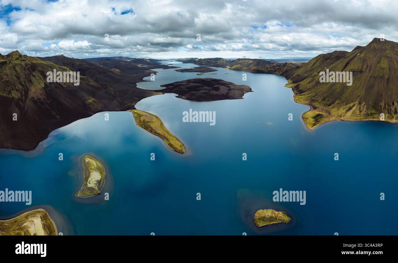 Vista aerea delle acque blu che riflettono il cielo nuvoloso e le aspre e scure montagne circostanti con macchie di verde, l'Islanda. Foto Stock