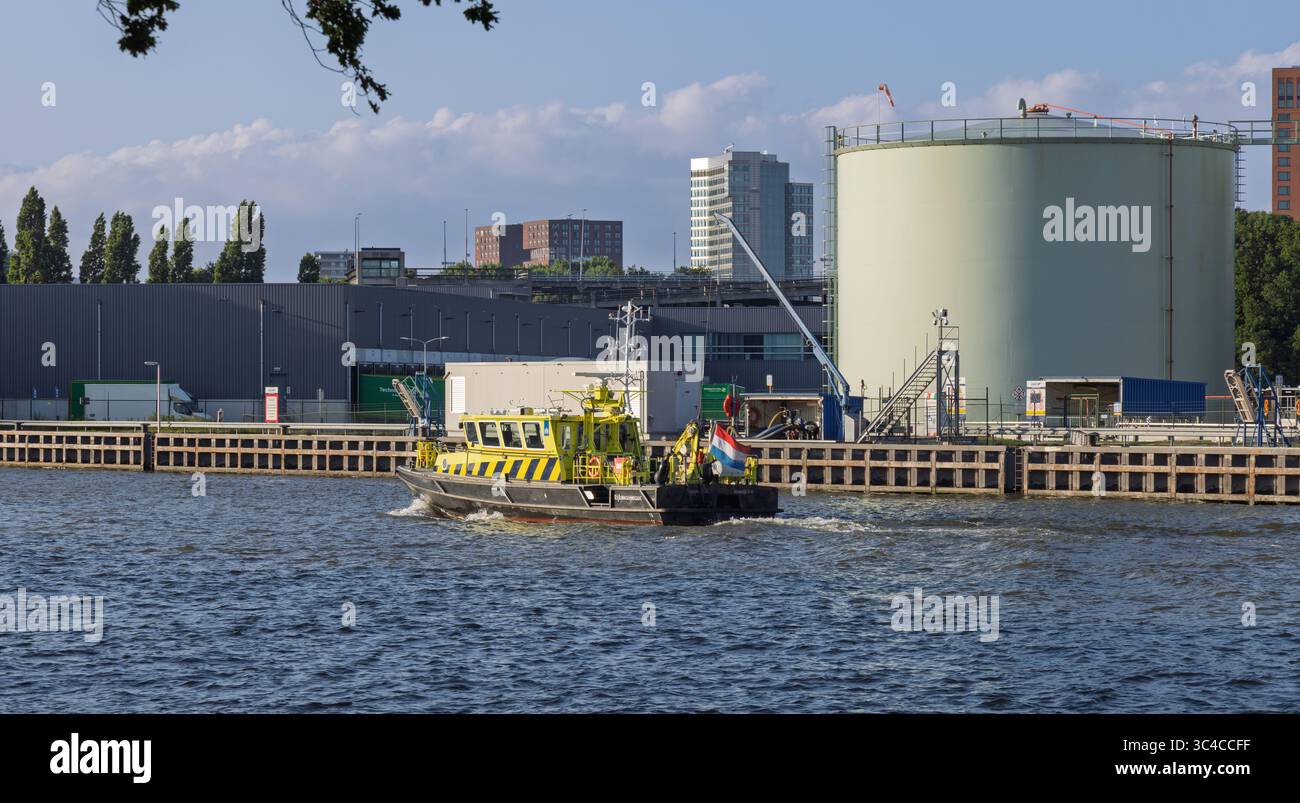La motovedetta rijkswaterstaat con bandiera olandese naviga lungo un corso d'acqua, passando attraverso grandi serbatoi industriali ed edifici urbani sotto un cielo limpido Foto Stock