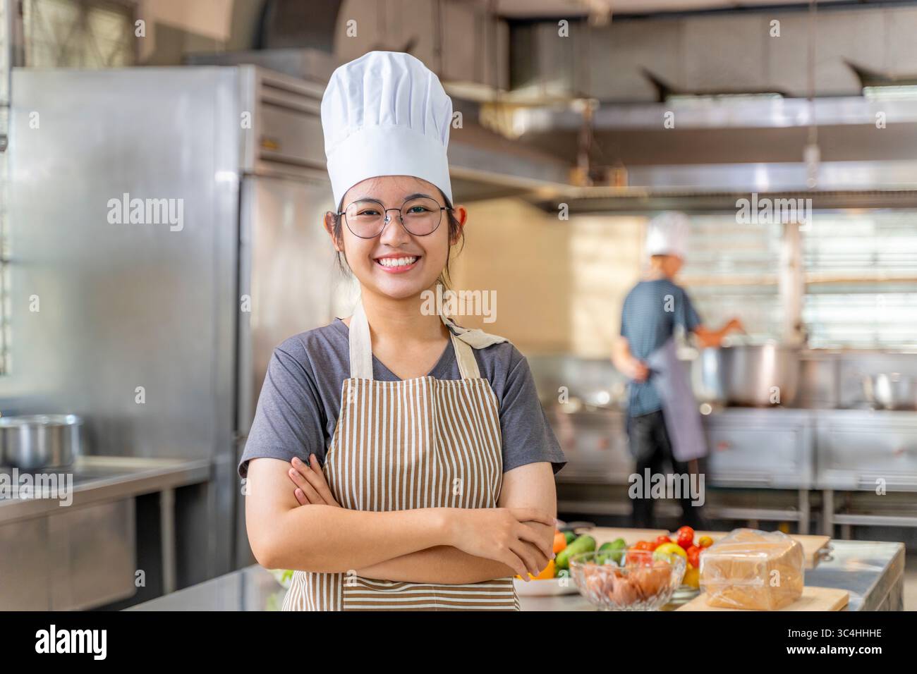 Giovane chef donna sicura di sé sorridente e in piedi con le braccia incrociate in una cucina commerciale. Indossa un tradizionale cappello da chef e grembiule a righe, con f Foto Stock