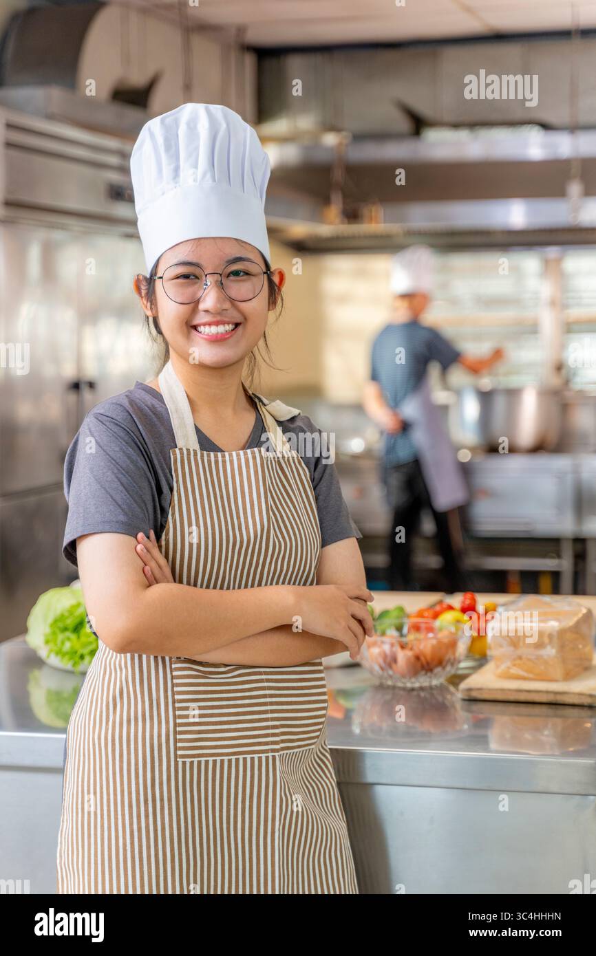 Giovane chef donna sicura di sé sorridente e in piedi con le braccia incrociate in una cucina commerciale. Indossa un tradizionale cappello da chef e grembiule a righe, con f Foto Stock