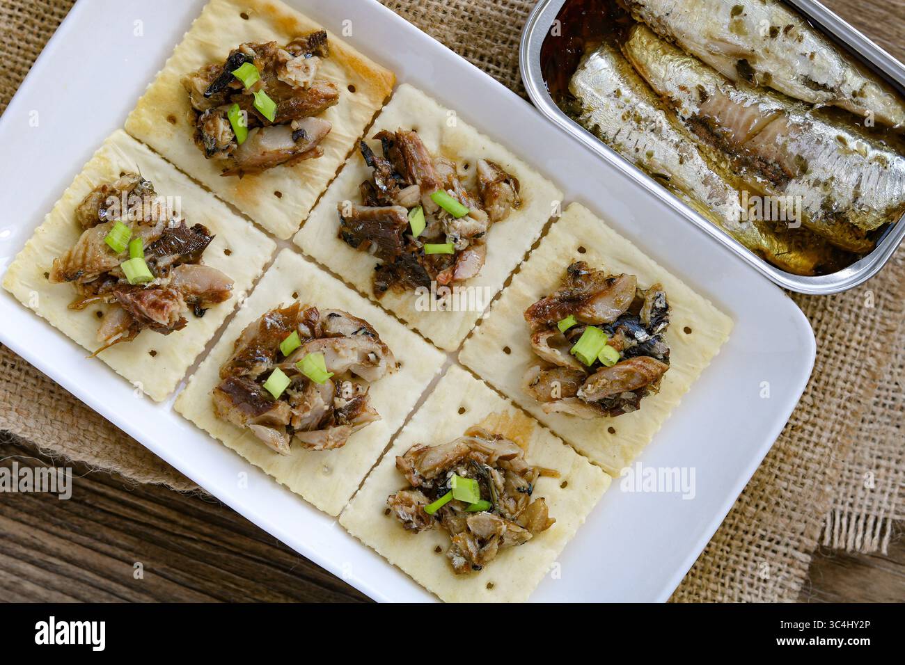 Un piatto di croccanti cracker quadrati, ciascuno ricoperto di sardine e una spolverata di cipolle verdi tritate, disposti su un piatto bianco rettangolare. Foto Stock