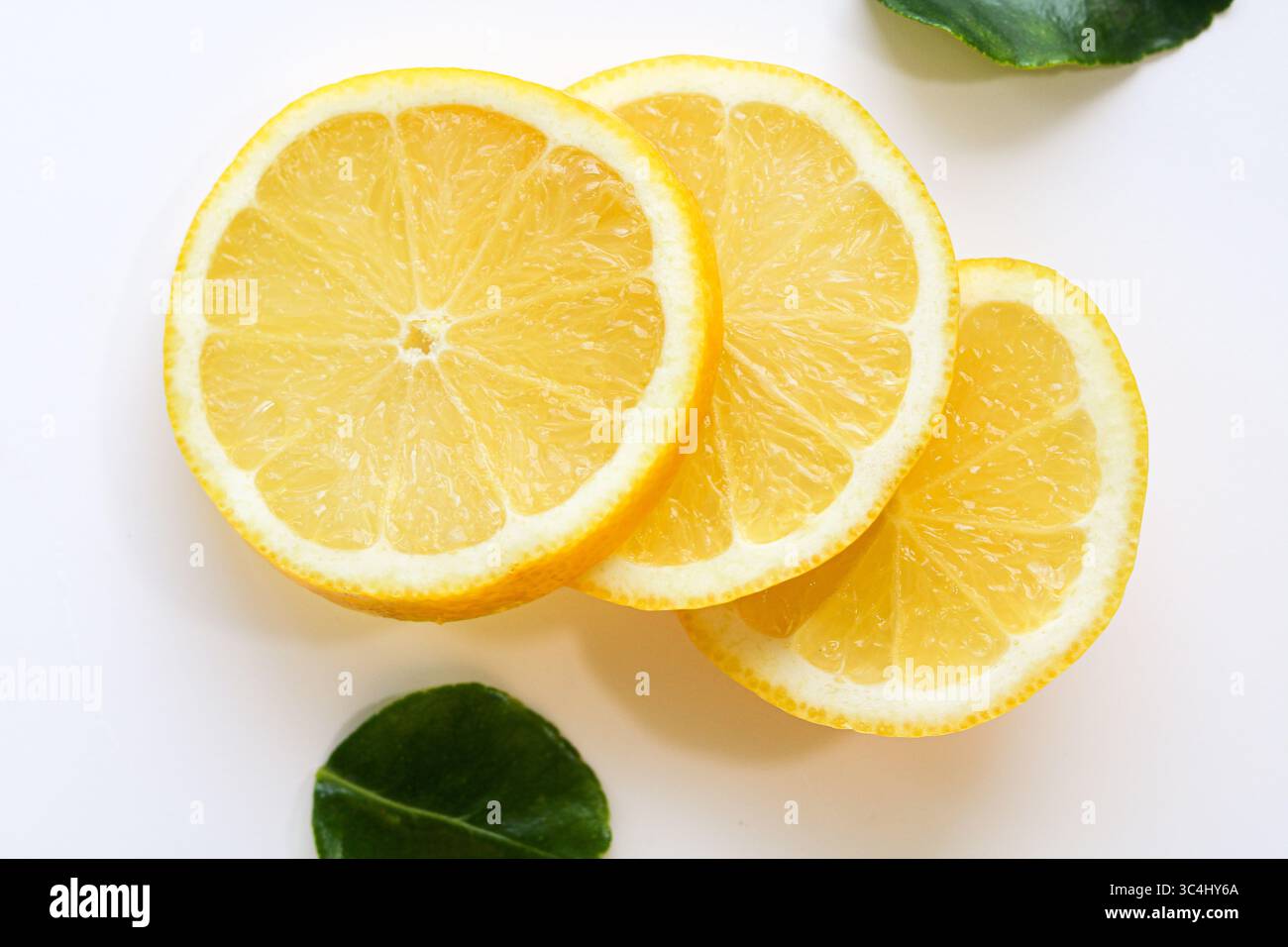 Tre fette luminose e succose di limone fresco, disposte ad arte su uno sfondo bianco pulito con foglie verdi sparse, pronte per bere o guarnire. Foto Stock