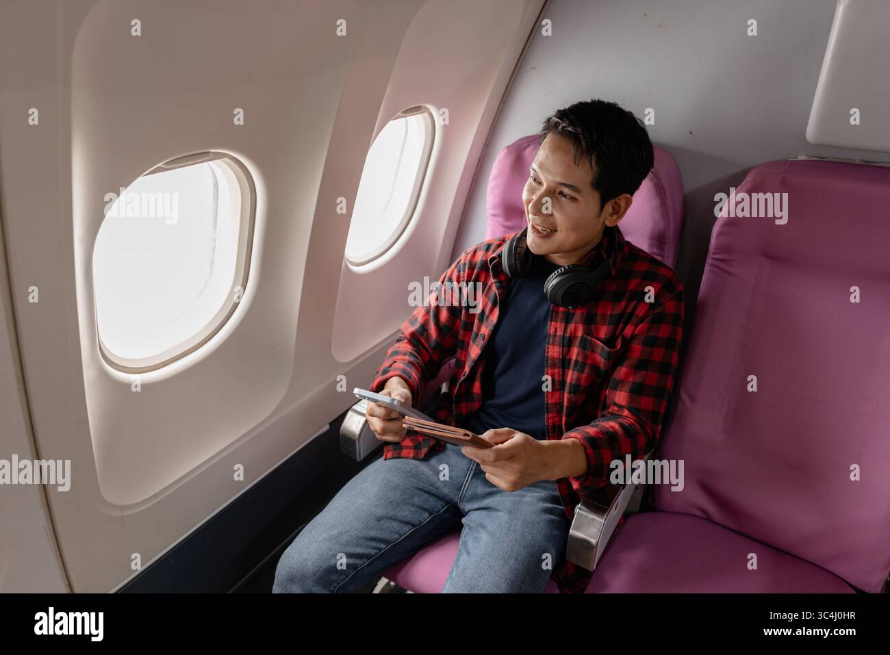 Un giovane uomo siede su un aereo, godendo il volo mentre controlla il suo smartphone e il passaporto. Sembra rilassato e contento, riflettendo il moderno Foto Stock