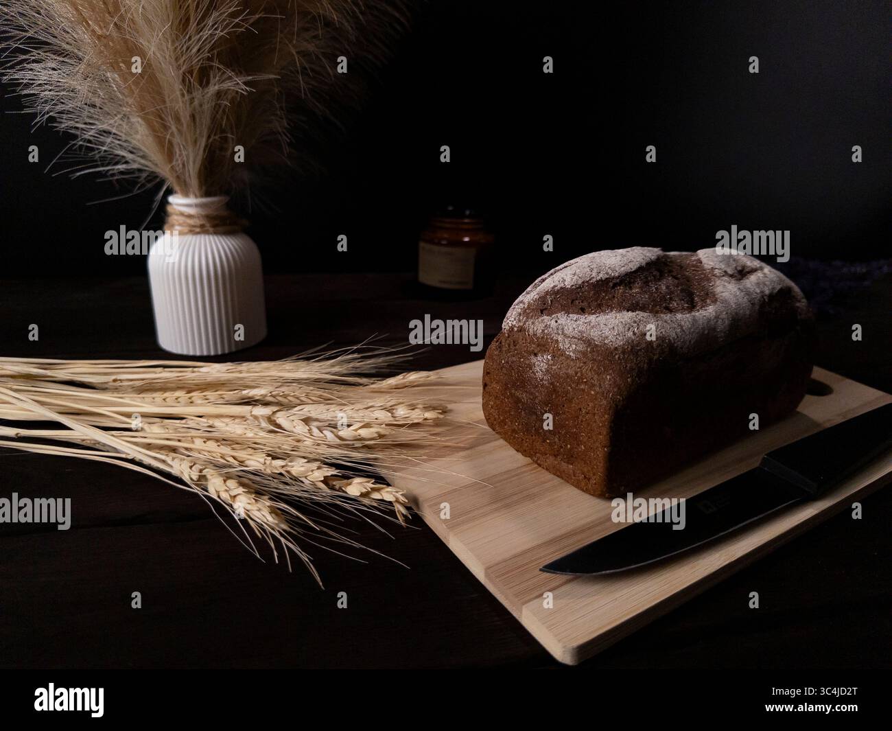 Una pagnotta rustica di pane su un tagliere di legno, accompagnata da gambi di grano essiccato e un vaso decorativo Foto Stock