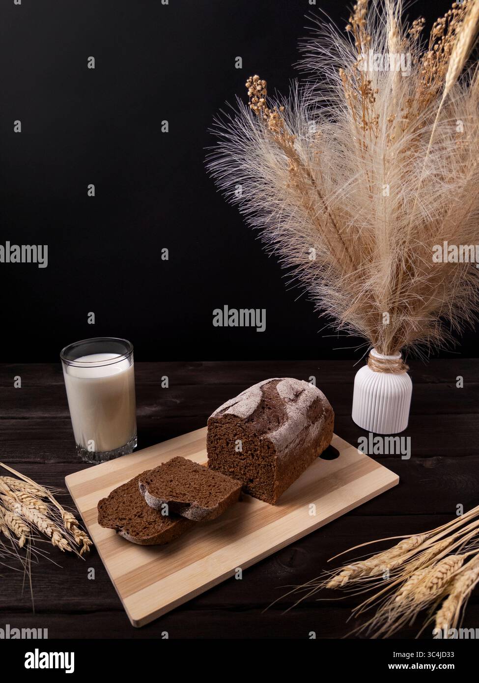 Pane rustico con fette su una tavola di legno, servito con un bicchiere di latte, grano e un vaso di fiori secchi su sfondo scuro Foto Stock