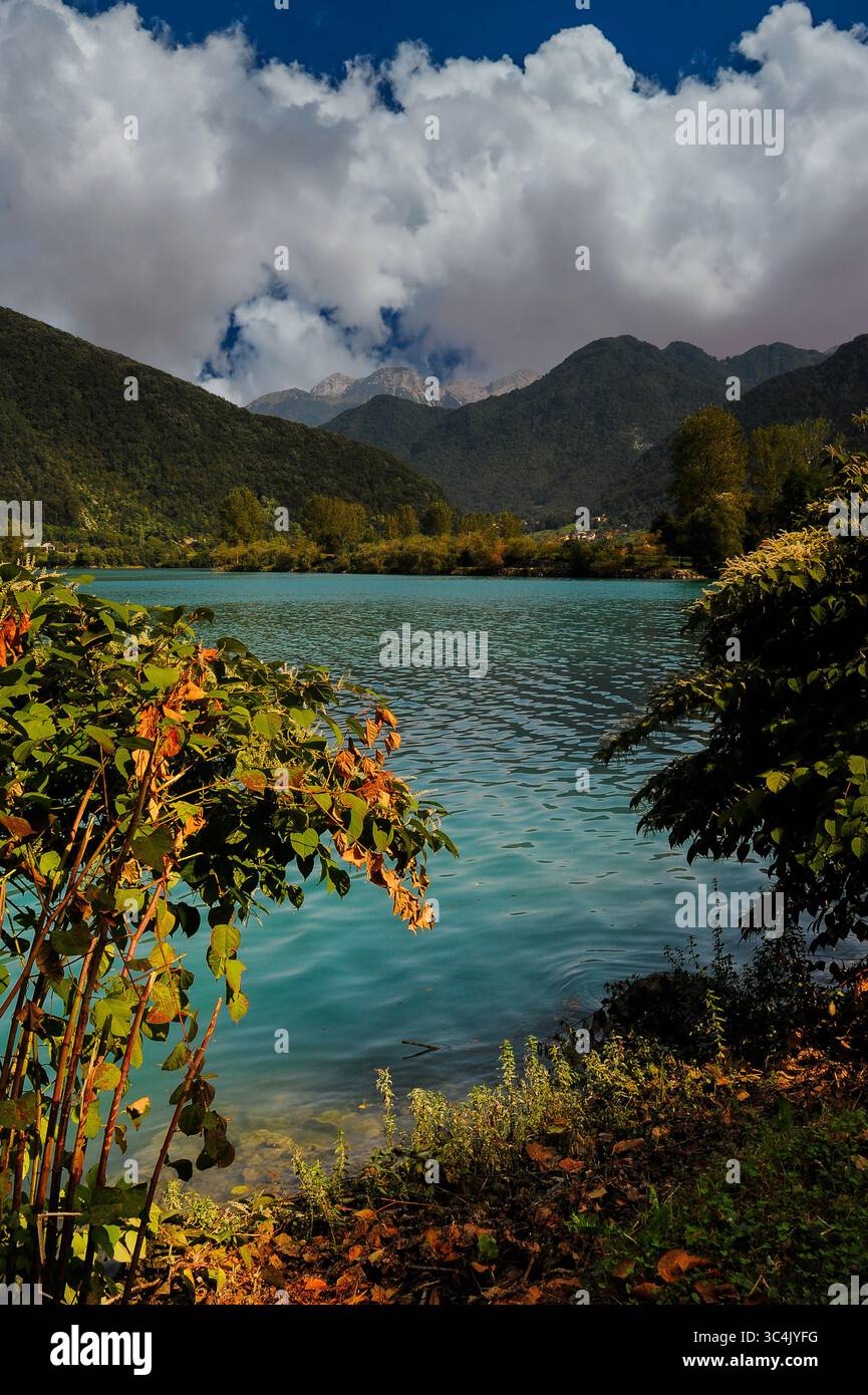 Le foglie dorate dell'inizio dell'autunno contrastano con le acque verdi smeraldo del bacino idrico di Soci, nella regione litorale della Slovenia occidentale. Vista sul lago all'inizio di settembre con nuvole che si ammassano sulle colline ricoperte di alberi sotto le vette delle Alpi Giulie nel Triglav o nel Parco Nazionale di Triglavski. Foto Stock