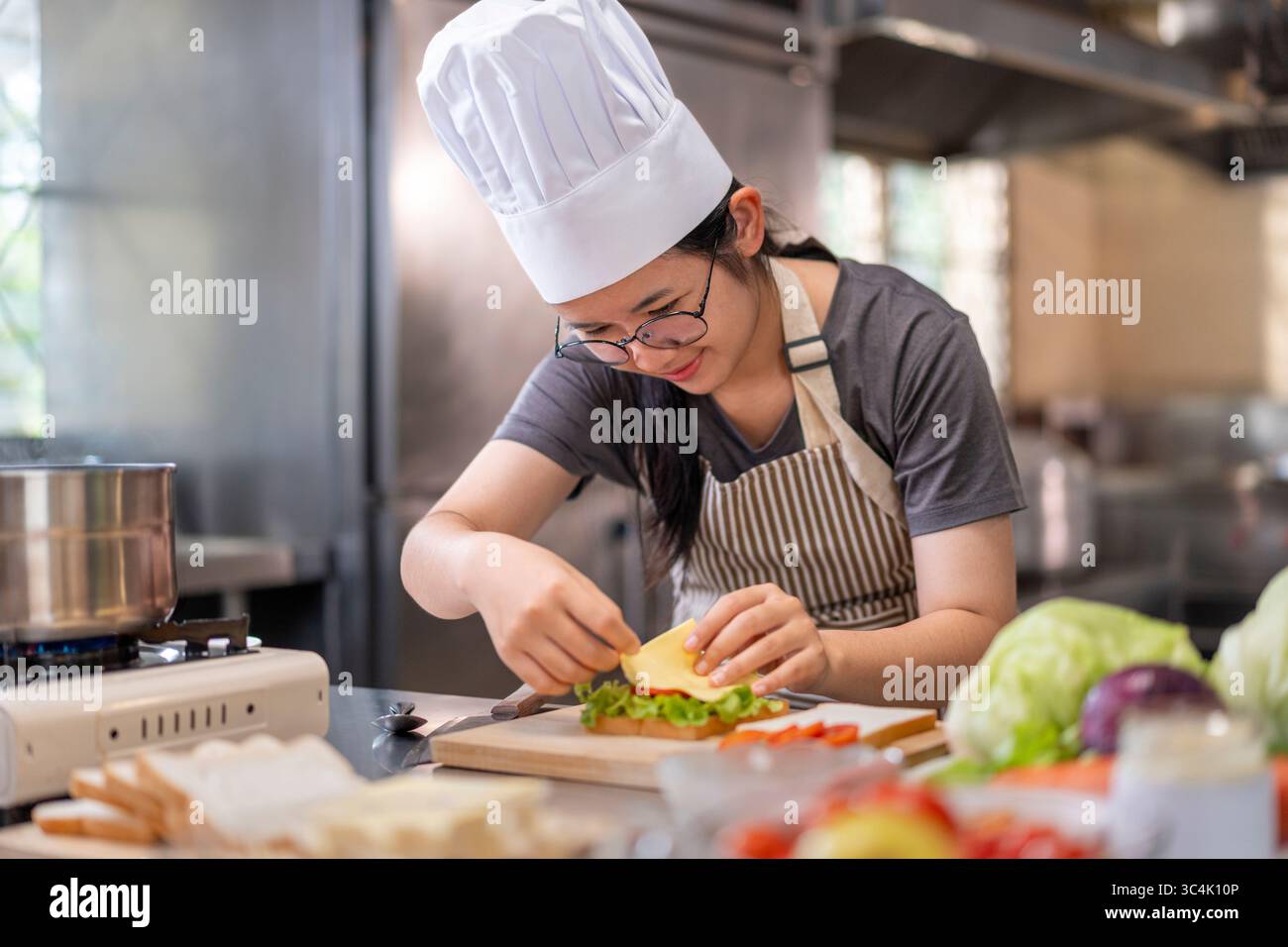 Una giovane e felice chef asiatica in una cucina professionale, che indossa un cappello da chef, sorride mentre assembla meticolosamente un panino fresco e salutare. Foto Stock