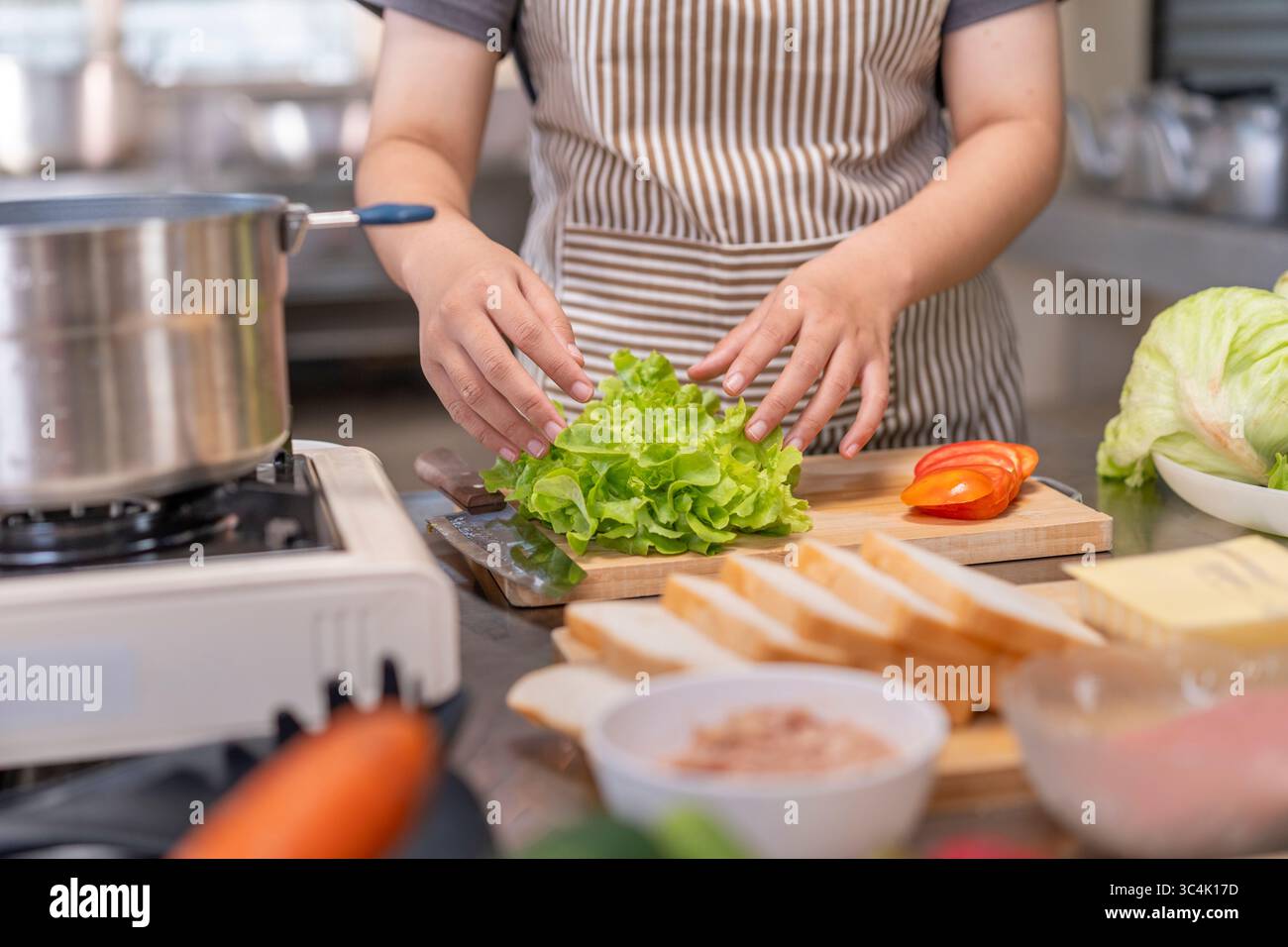 Una cuoca donna in grembiule prepara con cura lattuga verde fresca su un tagliere di legno con altri ingredienti. Ottimo per mangiare sano, preparazione alimentare Foto Stock