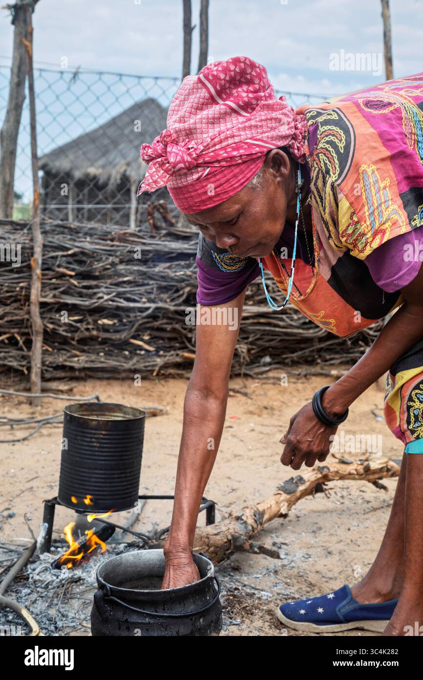 Villaggio rurale, sorridente vecchia africana che cucina all'aperto sopra un caminetto all'aperto. Capanne con tetto in paglia e recinzioni in legno sullo sfondo, Foto Stock