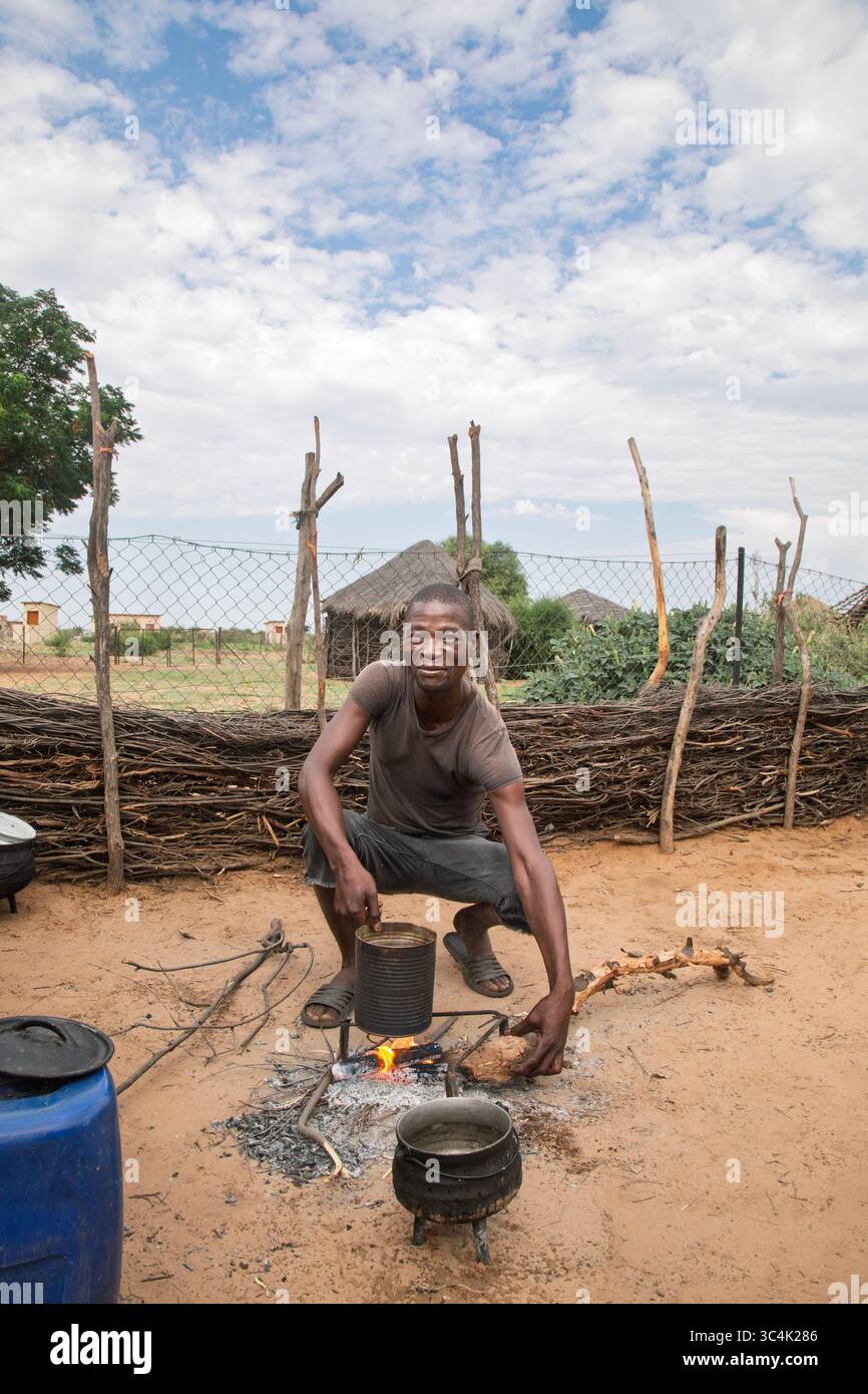 Villaggio rurale, sorridente uomo africano che cucina all'aperto su una cucina all'aperto con caminetto. Capanne con tetto in paglia e recinzione in legno sullo sfondo, spettacolo Foto Stock