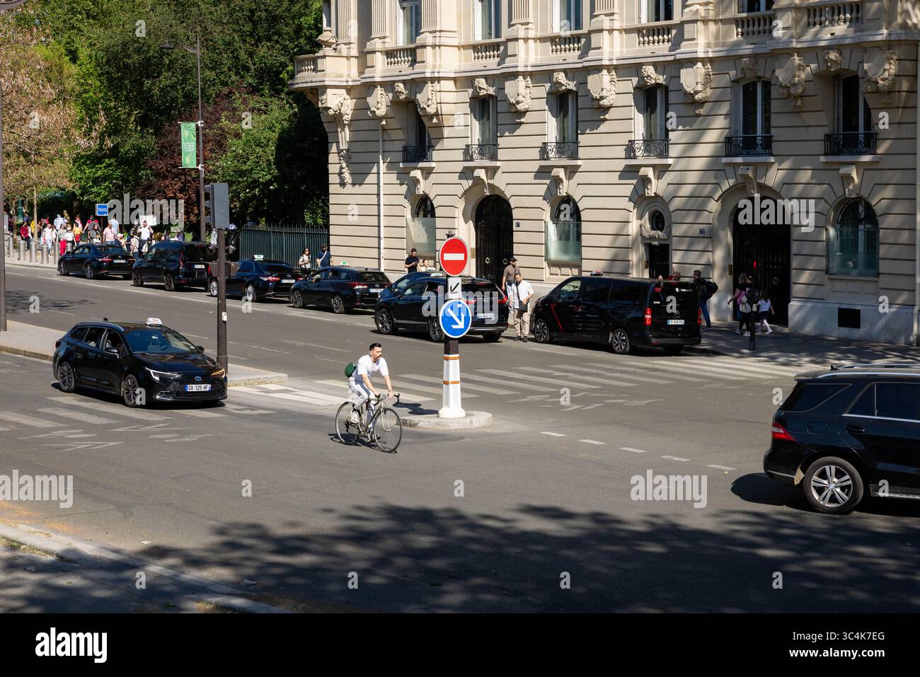 Una scena di strada parigina: Un ciclista bianco passa davanti a taxi neri e veicoli di lusso vicino a un grande edificio. La luce del sole proietta ombre. Ideale per t Foto Stock