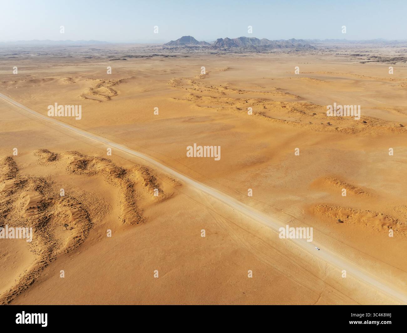 Veduta aerea di un paesaggio arido e soleggiato dove la strada pallida taglia le dune ocra verso montagne lontane, deserto del Namib, Namibia. Foto Stock