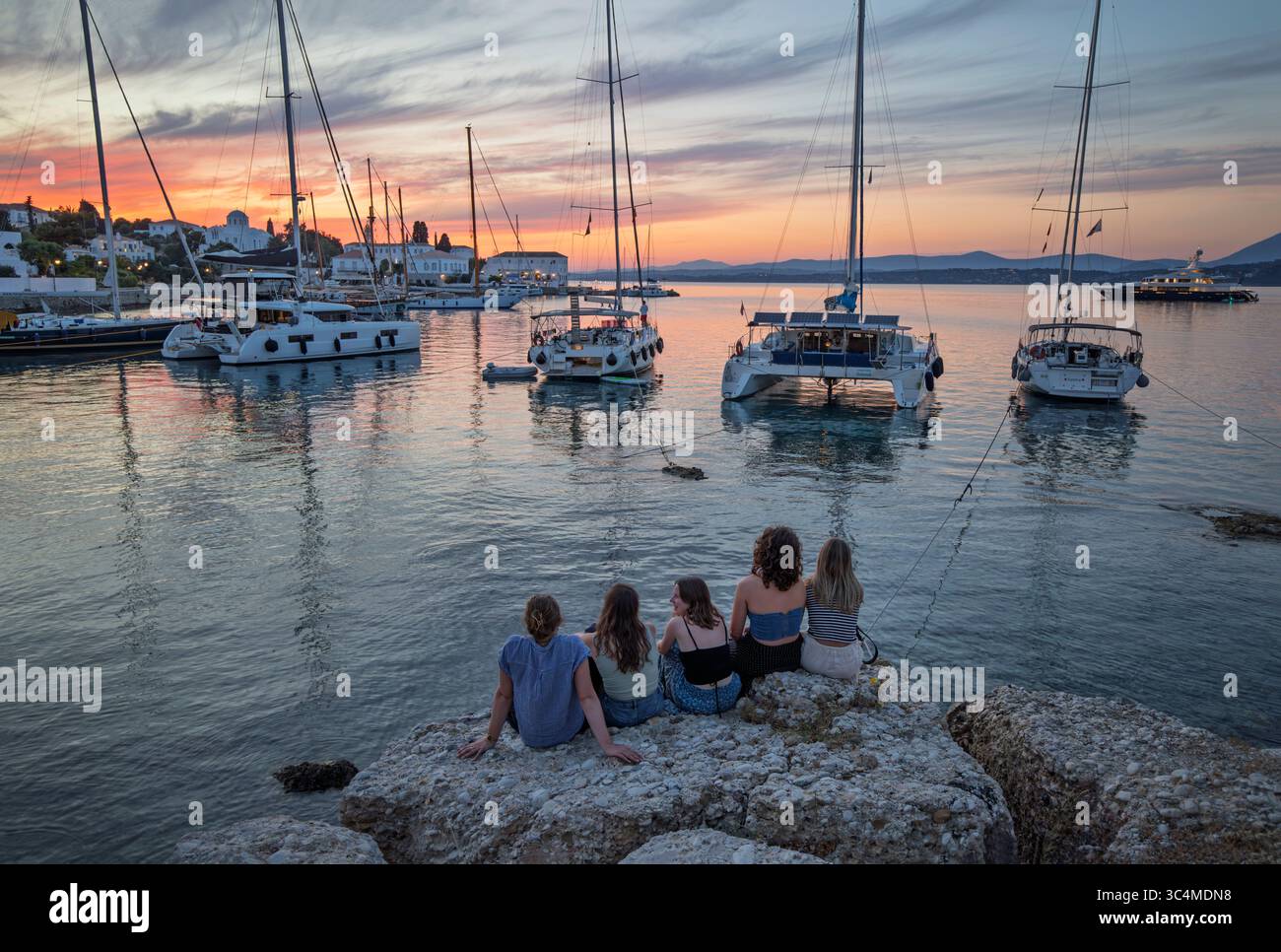 Gruppo di giovani seduti a guardare il tramonto sugli yacht nel vecchio porto di Palio Limani nella città di Spetses, Spetses, Isole Saroniche, Isole greche Foto Stock