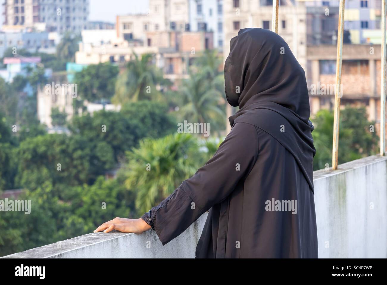 Donna musulmana bengalese in hijab nero su un tetto, in piedi da sola, che guarda in lontananza. Solitudine e contemplazione da una vista laterale. Foto Stock