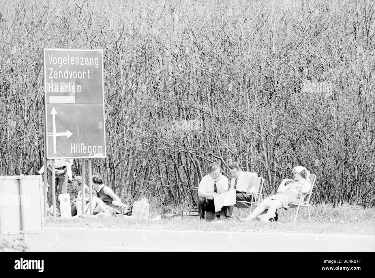Turismo su strada a Zilk, Turismo, turisti, 07-05-1970 Whizgle News, Dutch Desk, i Paesi Bassi, 1950-2000 Un gruppo di persone si rilassa lungo la strada, seduto su sedie a sdraio e impegnato in varie attività, con un cartello che indica le città vicine e delimitato da fitte foglie. Foto Stock