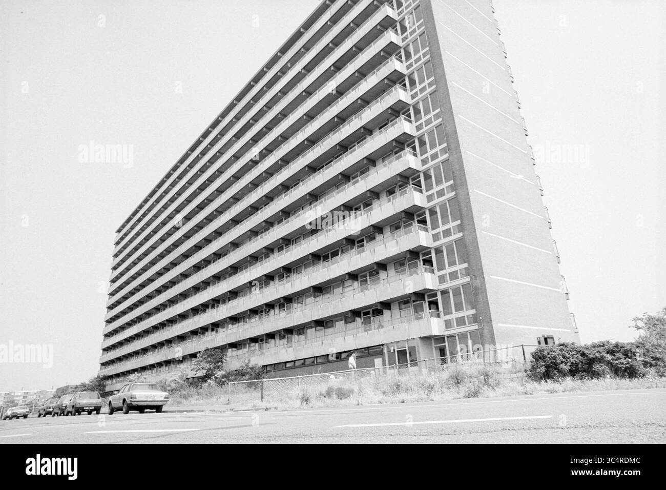 Flat Kruisberglaan Zeewijk IJmuiden, Flats, IJmuiden, Kruisberglaan, Paesi Bassi, 26-06-1989 Whizgle News, Dutch Desk, The Netherlands, 1950-2000 Un alto edificio residenziale angolare con balconi multipli e grandi finestre, adagiato su uno sfondo di paesaggi sparsi e una strada costeggiata da auto. L'architettura enfatizza un design moderno con una prospettiva leggermente inclinata, creando un impatto visivo sorprendente. Foto Stock