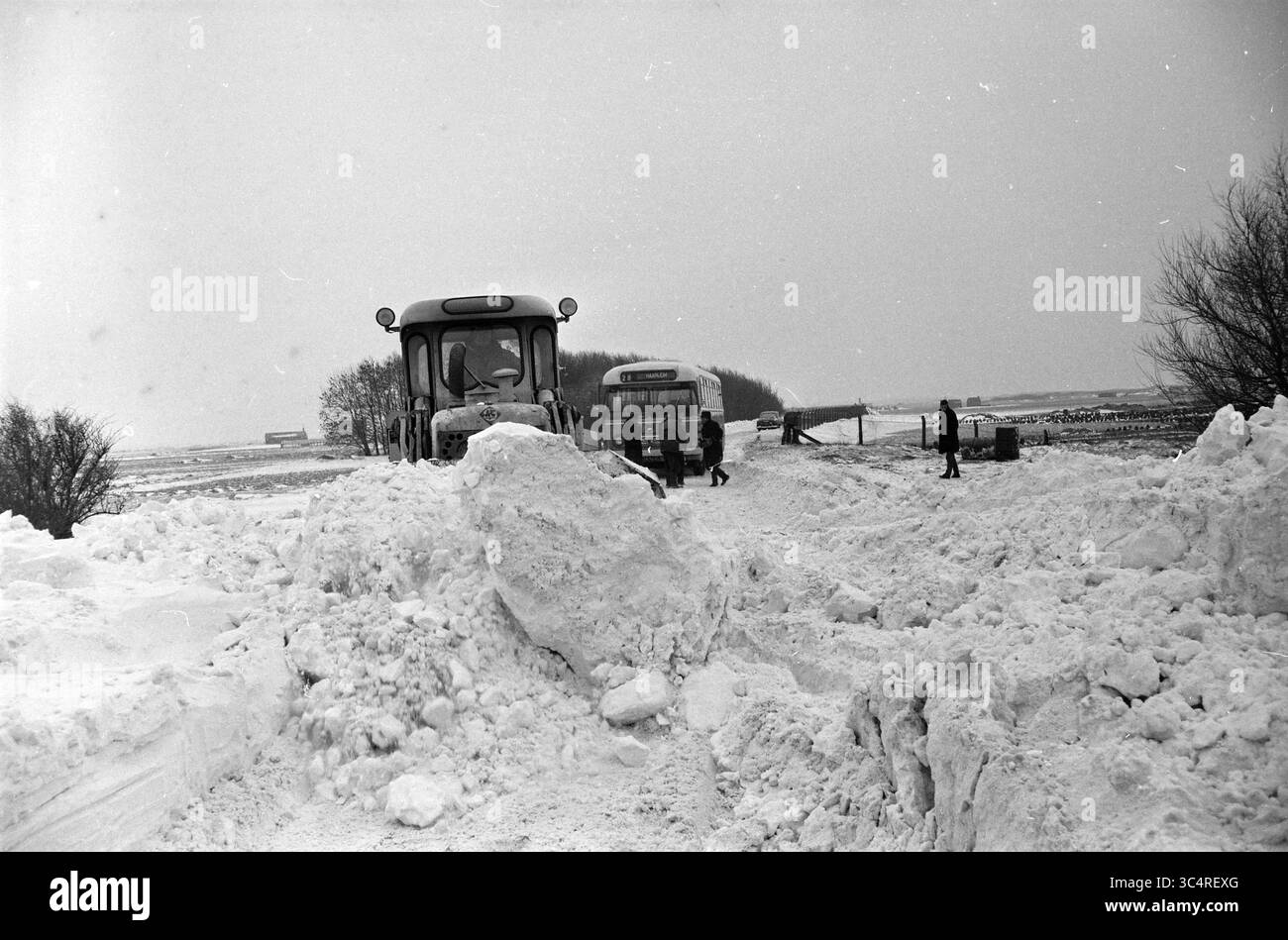 Pulizia della neve da strada/diga, 02-01-1963 Whizgle News, Dutch Desk, Paesi Bassi, 1950-2000 Un paesaggio innevato presenta una grande sponda di neve con due trattori e diverse persone che lavorano nelle vicinanze, navigando nelle condizioni invernali. Sullo sfondo, una linea sparsa di alberi e strutture lontane sono leggermente visibili, evidenziando la stanchezza della scena. Foto Stock