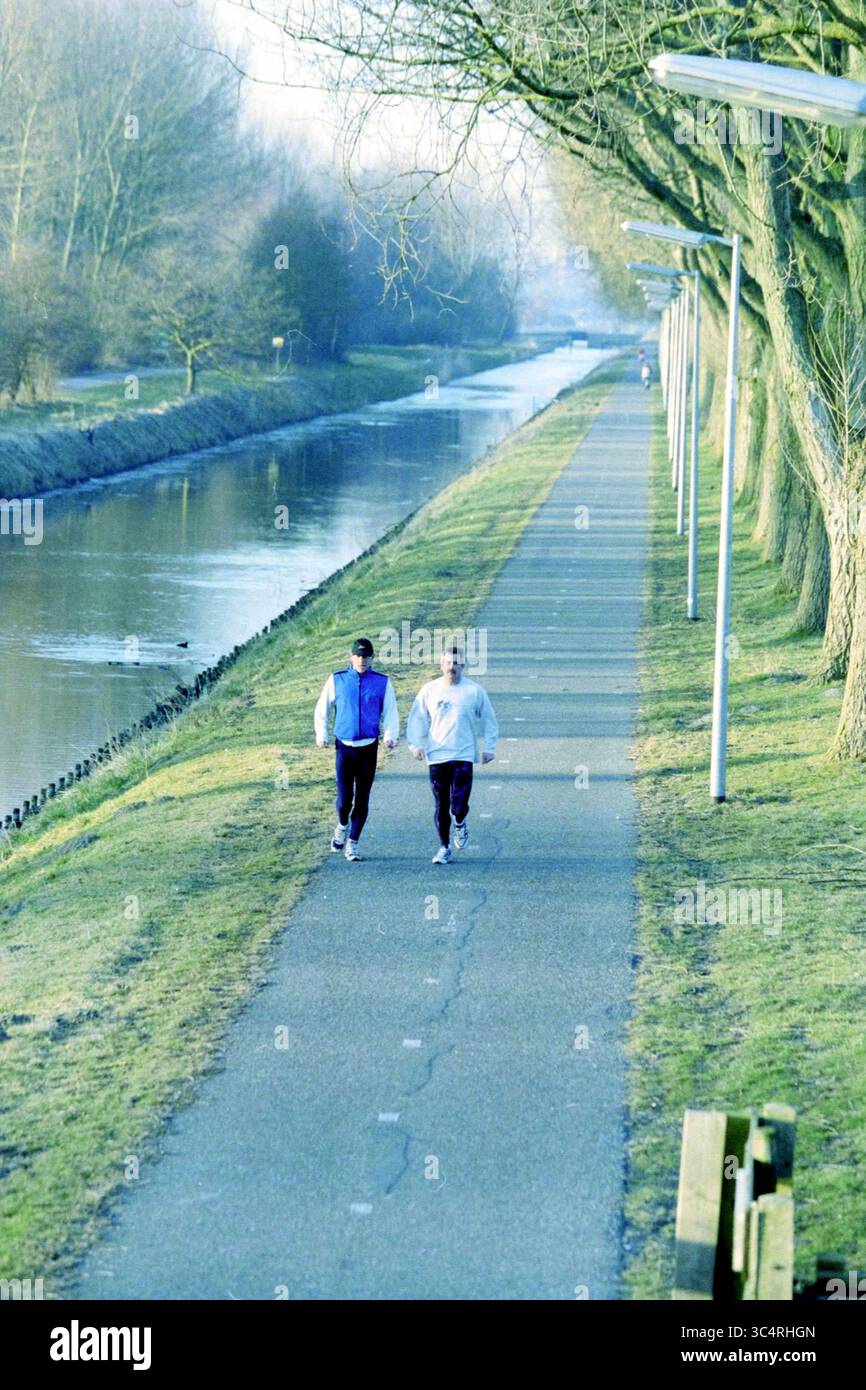 Jogger, Geniedijk, Hoofddorp, Geniedijk, Paesi Bassi, 24-02-2003 Whizgle News, Dutch Desk, Paesi Bassi, 1950-2000 due persone fanno jogging fianco a fianco lungo un tranquillo sentiero del canale, delimitato da alberi e fiancheggiato da lampioni. L'atmosfera serena suggerisce una mattinata tranquilla, con il sentiero che porta in lontananza. Foto Stock