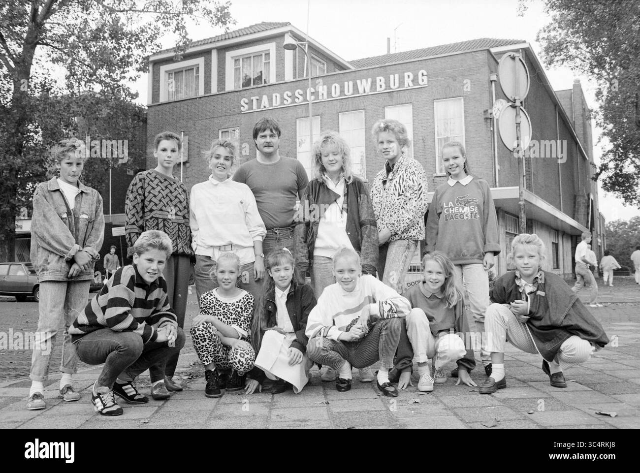 Gruppo di cabaret per bambini, IJmuiden, Cabaret, IJmuiden, Paesi Bassi, 18-09-1987 Whizgle News, Dutch Desk, Paesi Bassi, 1950-2000 Un gruppo di bambini e adolescenti posa di fronte a un edificio etichettato "Stadsschouwburg", che mostra un mix di stili di moda dell'epoca. La scena cattura un momento di cameratismo e gioia giovanile, sullo sfondo del paesaggio urbano. Foto Stock