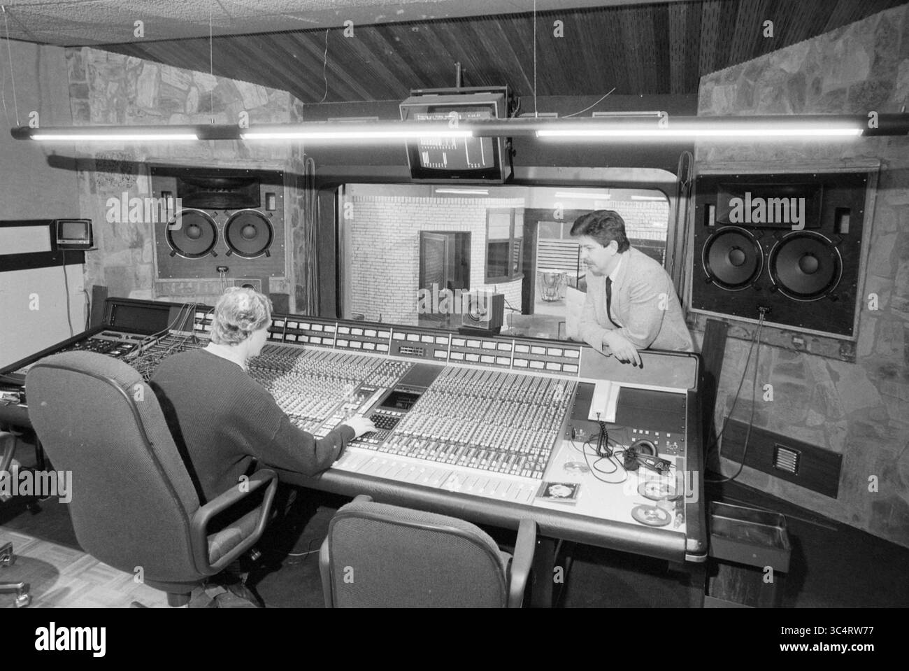 Studio di registrazione EMI Bovema, Heemstede, Bronsteeweg, Paesi Bassi, 29-01-1987 Whizgle News, Dutch Desk, Paesi Bassi, 1950-2000 in un vivace studio di registrazione, un tecnico del suono regola abilmente i livelli su una grande console di mixaggio, mentre un collega osserva intensamente dalle vicinanze, impegnato nel processo creativo della produzione musicale. I grandi altoparlanti affiancano la stanza, sottolineando l'attenzione alla qualità audio. Foto Stock