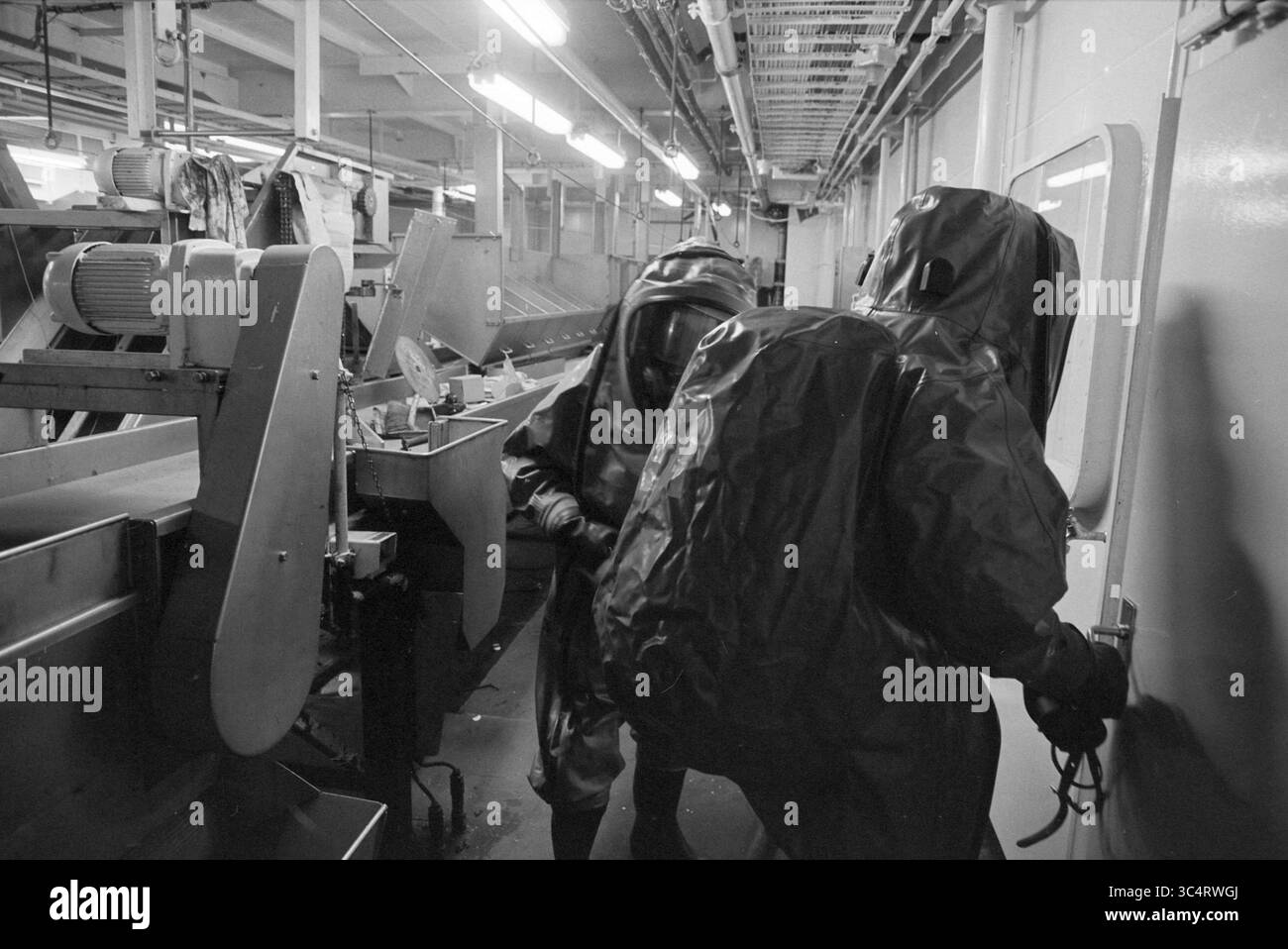 Esercitazione dei vigili del fuoco, Tridens, 22-03-1994 Whizgle News, Dutch Desk, Paesi Bassi, 1950-2000 due persone in tute protettive navigano in uno spazio industriale ristretto, concentrandosi su un apparato simile a un trasportatore. L'ambiente mostra segni di essere un'area di lavorazione, con macchinari e attrezzature che rivestono le pareti. Foto Stock
