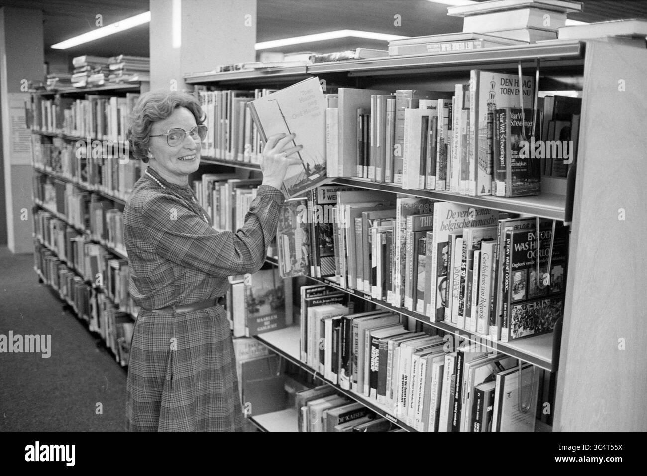 Library Whizgle News, Dutch Desk, Paesi Bassi, 1950-2000 Una donna con un vestito a quadri si erge accanto a un'alta libreria piena di libri, tenendone uno in mano e sorridente. Appare impegnata e attenta, circondata da numerosi titoli in un ambiente accogliente della biblioteca. Foto Stock